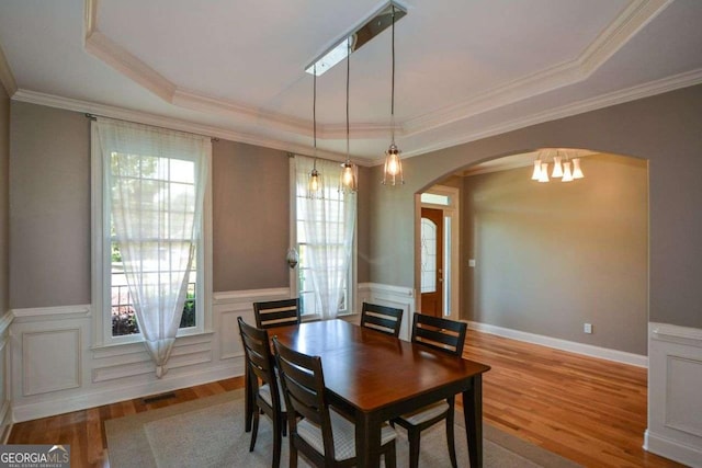dining space featuring arched walkways, plenty of natural light, a tray ceiling, wood finished floors, and wainscoting