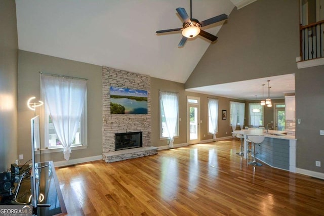 living area featuring a ceiling fan, baseboards, wood finished floors, a stone fireplace, and high vaulted ceiling