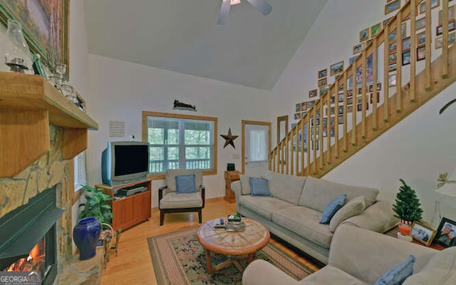 living area featuring high vaulted ceiling, a warm lit fireplace, stairway, a ceiling fan, and wood finished floors