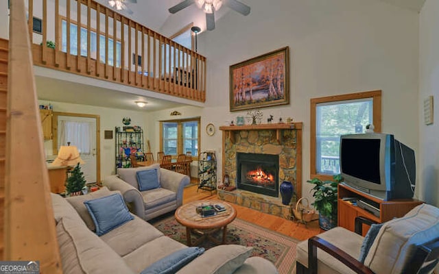 living room featuring a towering ceiling, a ceiling fan, plenty of natural light, a stone fireplace, and wood finished floors