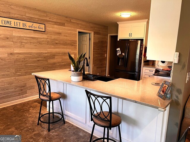 kitchen with black fridge with ice dispenser, wood walls, a breakfast bar, white cabinets, and a textured ceiling