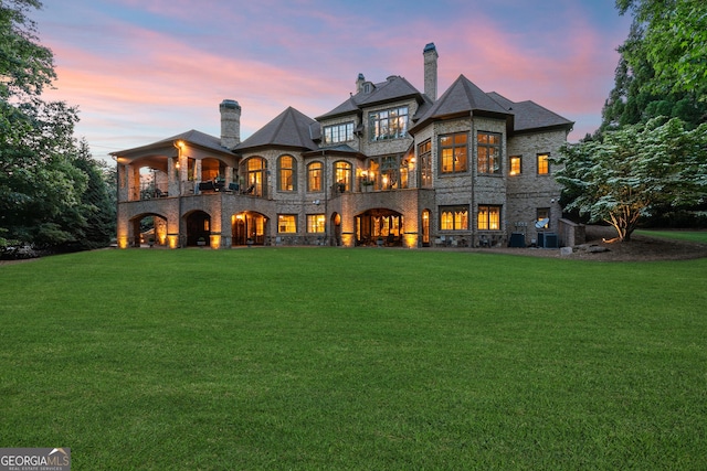 back of property at dusk with stone siding, a lawn, and a chimney