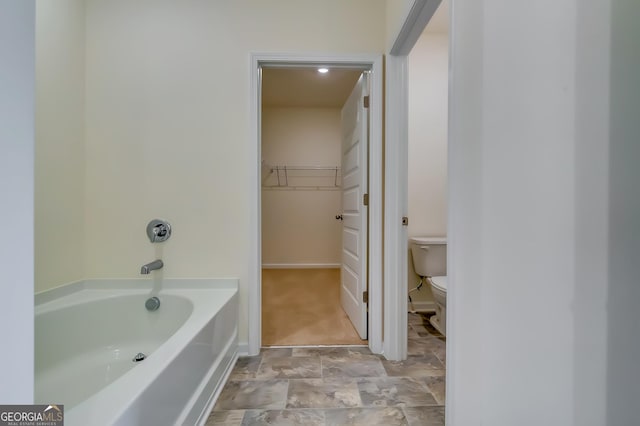 bathroom with toilet, a bath, a spacious closet, stone finish floors, and baseboards