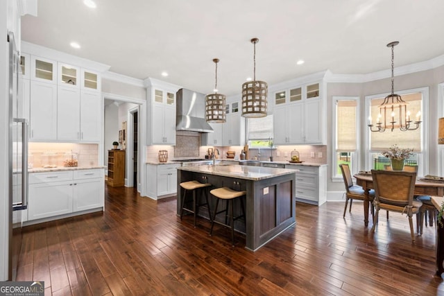 kitchen with wall chimney range hood, a chandelier, a center island with sink, crown molding, and white cabinetry