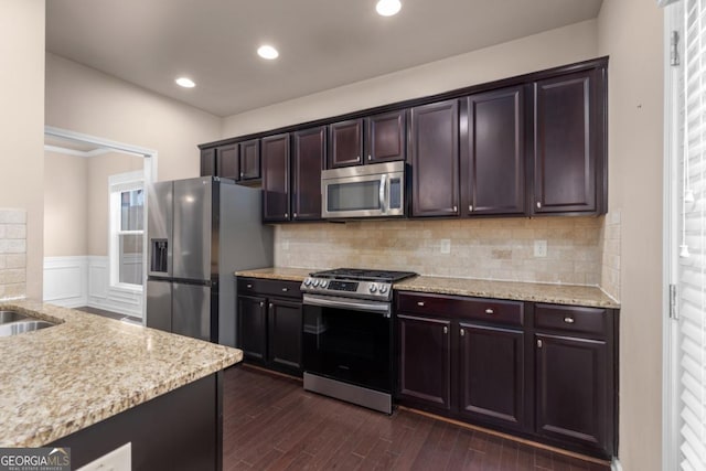 kitchen featuring stainless steel appliances, dark wood-style floors, light stone counters, dark brown cabinetry, and a wainscoted wall