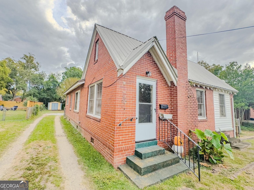 view of front of home featuring a chimney, entry steps, brick siding, fence, and a storage shed