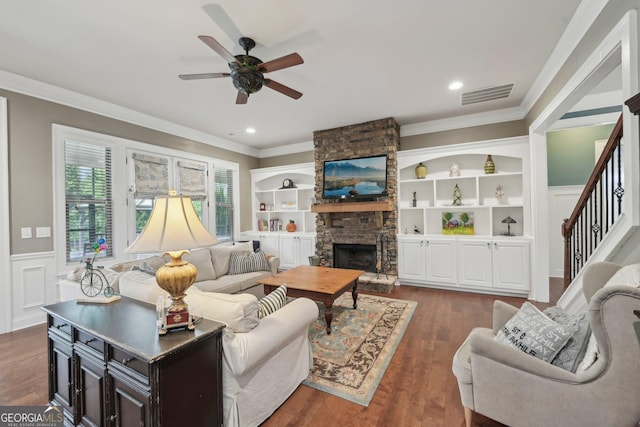 living room featuring ceiling fan, wainscoting, a stone fireplace, crown molding, and stairway
