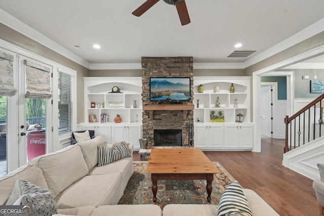 living room with crown molding, a ceiling fan, stairs, dark wood-style flooring, and a stone fireplace
