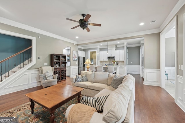 living area featuring ceiling fan, wood-type flooring, stairs, a decorative wall, and wainscoting