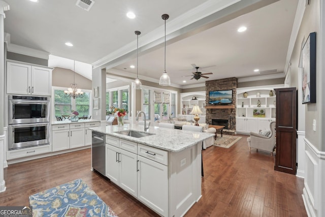 kitchen with a sink, stainless steel appliances, a ceiling fan, a stone fireplace, and white cabinetry