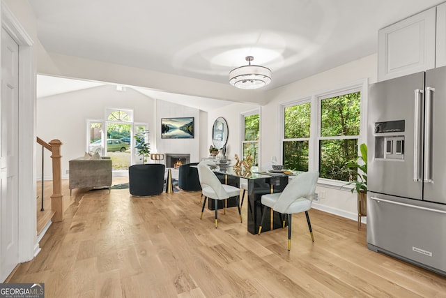 dining space featuring a warm lit fireplace, light wood finished floors, vaulted ceiling, and baseboards