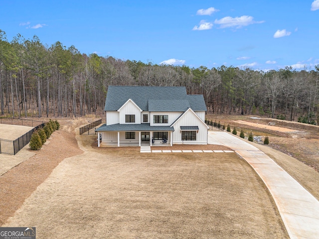 modern farmhouse featuring a view of trees, concrete driveway, covered porch, roof with shingles, and fence