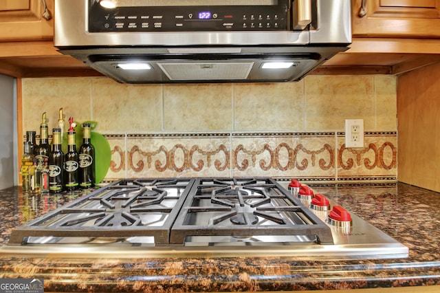 kitchen view of tasteful backsplash, exhaust hood, stainless steel gas stovetop, and dark stone counters