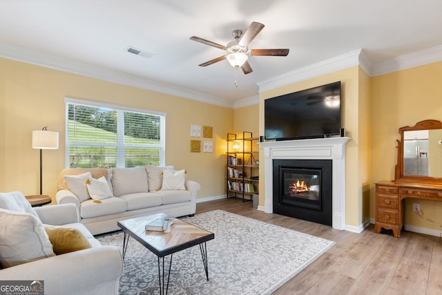 living room with ornamental molding, a ceiling fan, light wood-type flooring, a glass covered fireplace, and baseboards