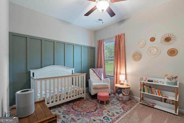 bedroom with a nursery area, a decorative wall, carpet, ceiling fan, and wainscoting