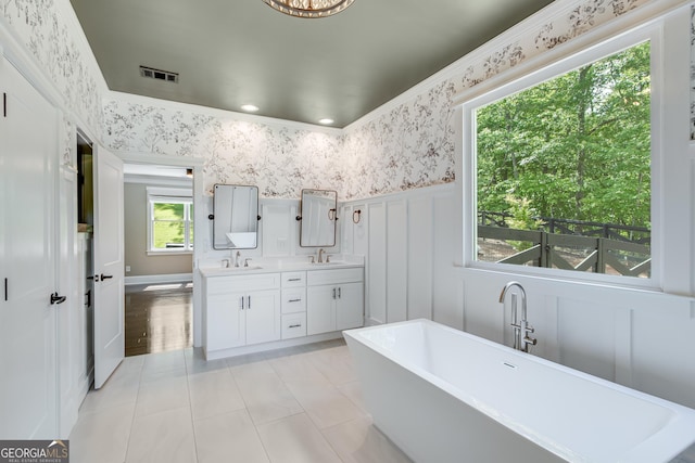 full bath featuring wallpapered walls, wainscoting, double vanity, a freestanding bath, and tile patterned flooring