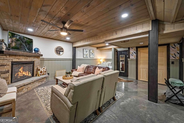 living area with finished concrete floors, wooden ceiling, a stone fireplace, wainscoting, and a ceiling fan