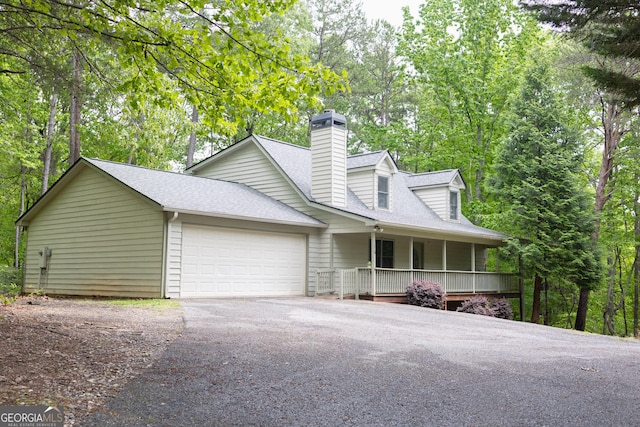 view of front facade featuring asphalt driveway, a shingled roof, an attached garage, covered porch, and a chimney