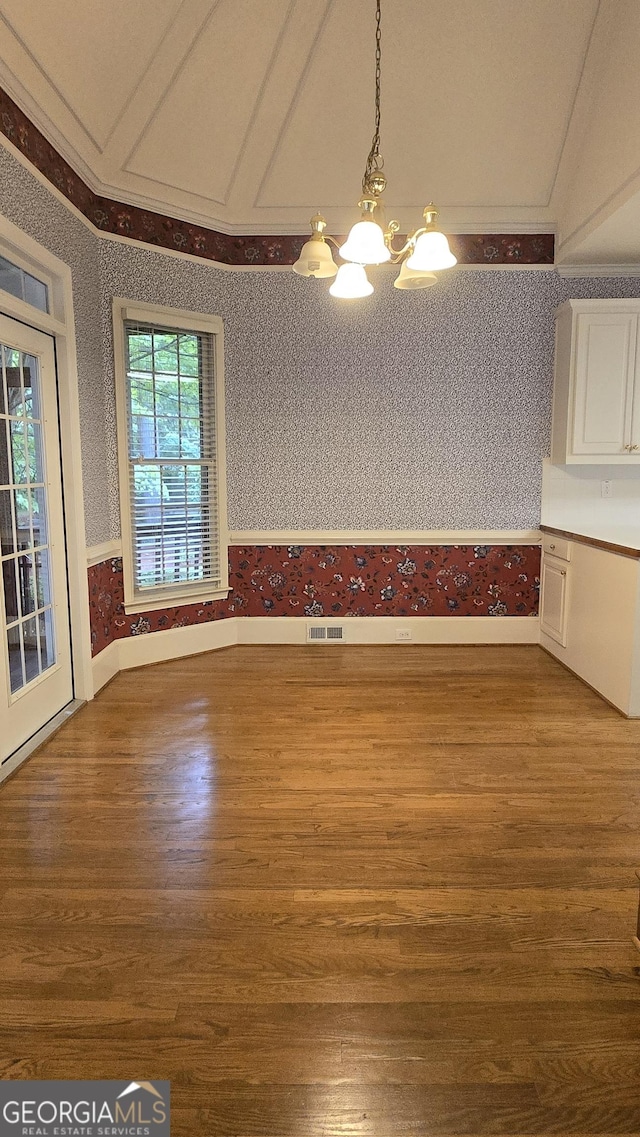 unfurnished dining area with a chandelier, wallpapered walls, wood finished floors, a wainscoted wall, and crown molding