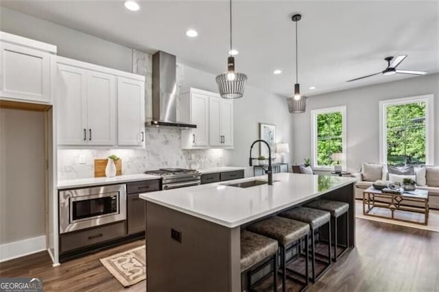 kitchen featuring wall chimney exhaust hood, a sink, appliances with stainless steel finishes, open floor plan, and dark wood-style flooring