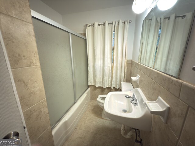 bathroom featuring bath / shower combo with glass door, tile walls, toilet, and a sink