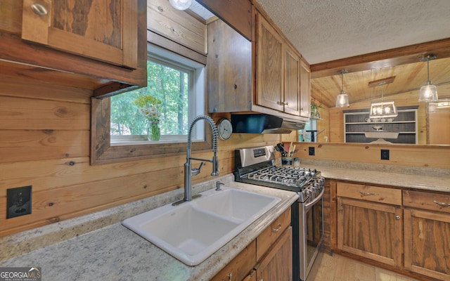 kitchen featuring stainless steel gas stove, a sink, under cabinet range hood, brown cabinets, and wood walls