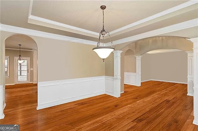 unfurnished dining area featuring arched walkways, ornate columns, a tray ceiling, wood finished floors, and wainscoting