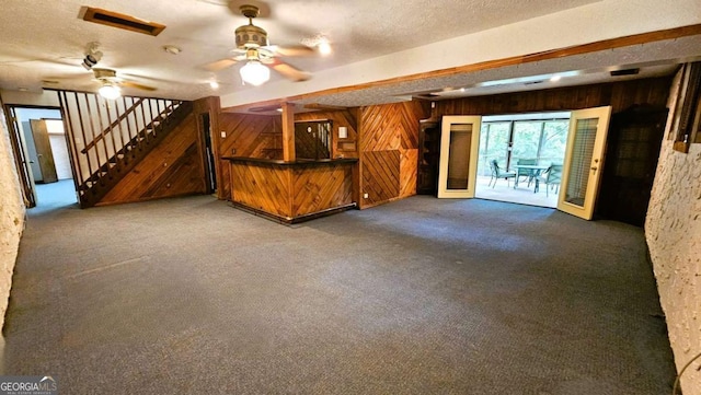 unfurnished living room with a dry bar, wood walls, carpet floors, a ceiling fan, and stairway
