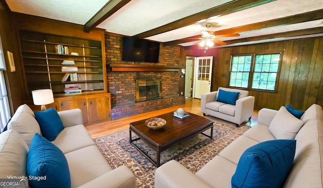 living area with beamed ceiling, ceiling fan, wood finished floors, wood walls, and a brick fireplace