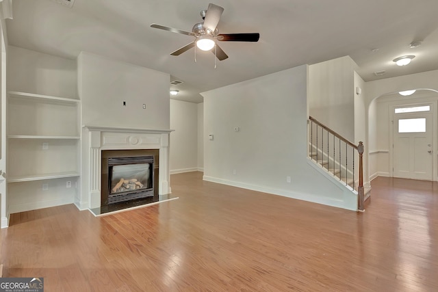 unfurnished living room with wood finished floors, a ceiling fan, stairway, a glass covered fireplace, and arched walkways
