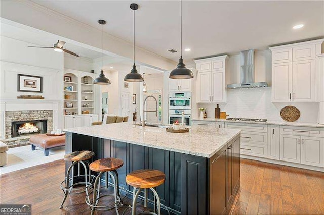 kitchen with wall chimney exhaust hood, appliances with stainless steel finishes, white cabinetry, ornamental molding, and a kitchen island with sink