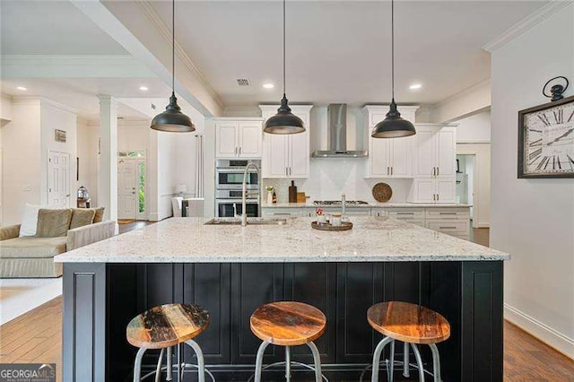 kitchen featuring wall chimney range hood, appliances with stainless steel finishes, white cabinetry, a large island, and ornamental molding