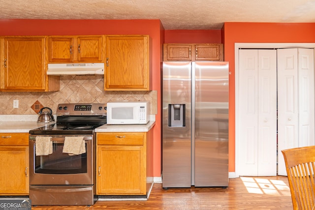 kitchen featuring stainless steel appliances, under cabinet range hood, light wood-style floors, light countertops, and tasteful backsplash