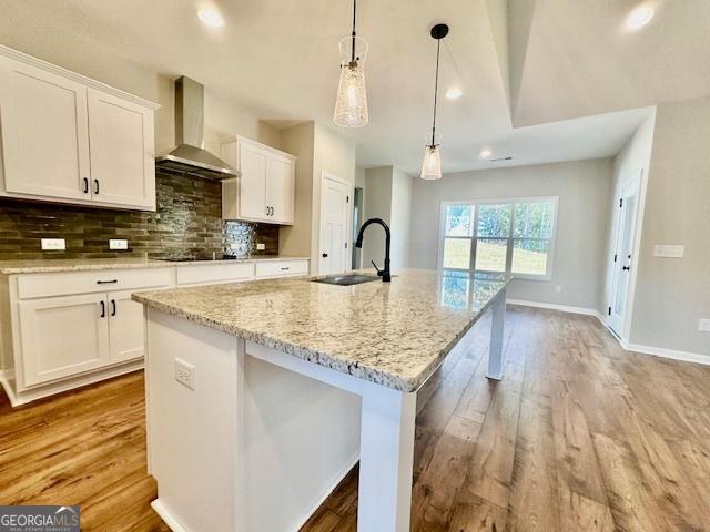 kitchen with wall chimney exhaust hood, a sink, a center island with sink, white cabinets, and backsplash