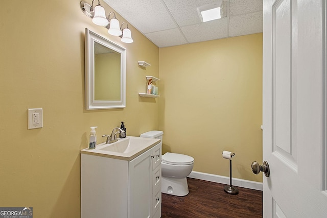 half bathroom featuring a drop ceiling, toilet, wood finished floors, vanity, and baseboards