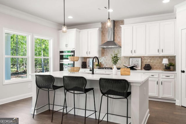 kitchen featuring wall chimney range hood, double oven, a kitchen island with sink, white cabinets, and tasteful backsplash