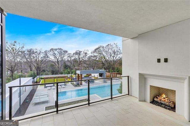 balcony featuring a warm lit fireplace, view of pool area, a patio, and view of scattered trees