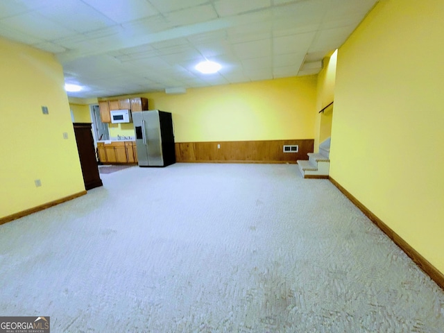unfurnished living room with light colored carpet, a wainscoted wall, stairway, a drop ceiling, and wood walls