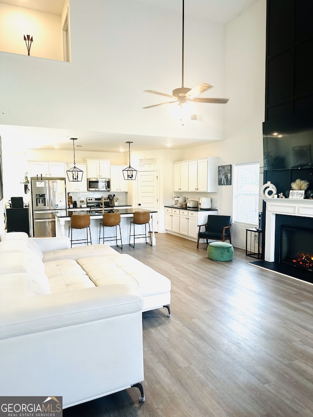 living room with a high ceiling, a ceiling fan, light wood-style flooring, and a lit fireplace