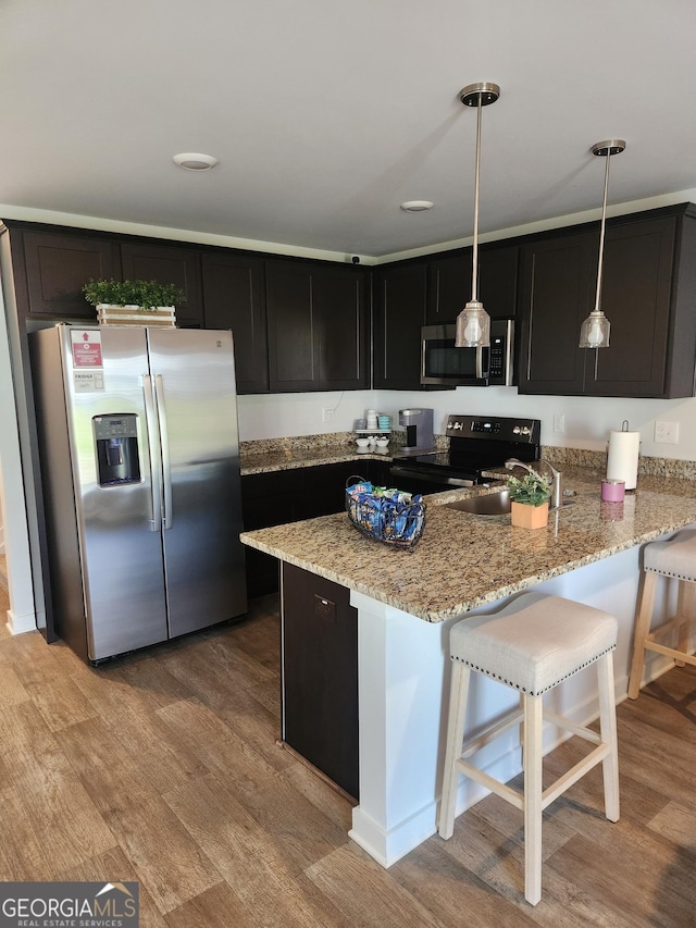 kitchen featuring appliances with stainless steel finishes, light wood finished floors, light stone counters, dark cabinetry, and a kitchen breakfast bar