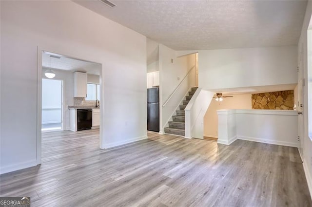 unfurnished living room featuring stairs, light wood-style floors, a textured ceiling, baseboards, and a ceiling fan