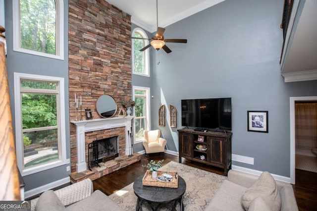 living area with a towering ceiling, crown molding, a stone fireplace, a ceiling fan, and wood finished floors