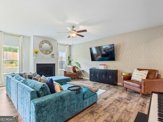 living room featuring wallpapered walls, wood finished floors, healthy amount of natural light, a glass covered fireplace, and ceiling fan