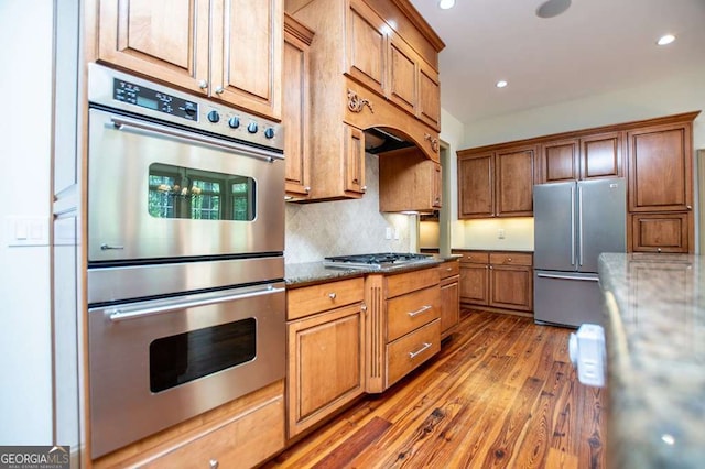 kitchen featuring stainless steel appliances, wood finished floors, backsplash, dark stone countertops, and brown cabinets