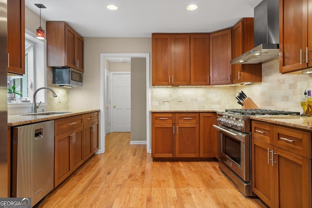 kitchen featuring stainless steel appliances, wall chimney range hood, a sink, light wood finished floors, and light stone countertops