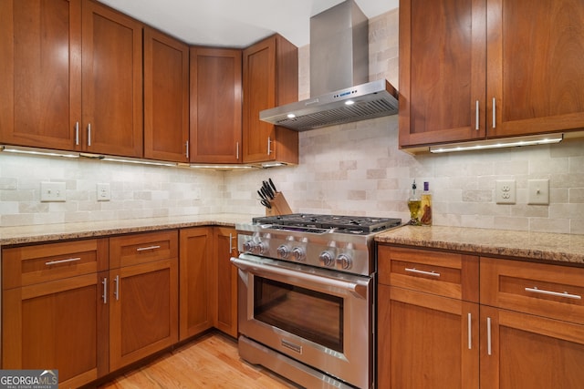 kitchen featuring stainless steel range, wall chimney exhaust hood, brown cabinets, and light stone counters