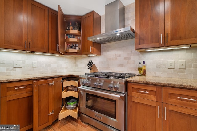kitchen featuring stainless steel stove, wall chimney exhaust hood, light stone countertops, tasteful backsplash, and brown cabinets