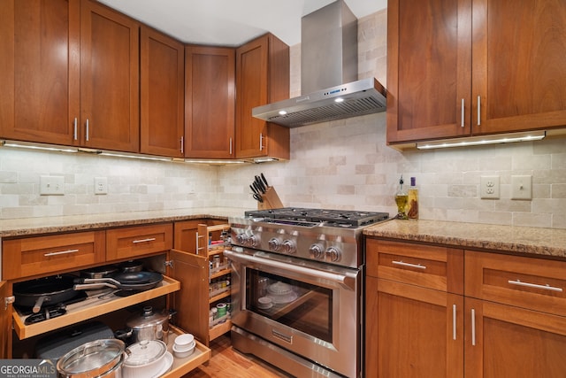 kitchen with high end range, wall chimney range hood, light stone countertops, brown cabinets, and decorative backsplash