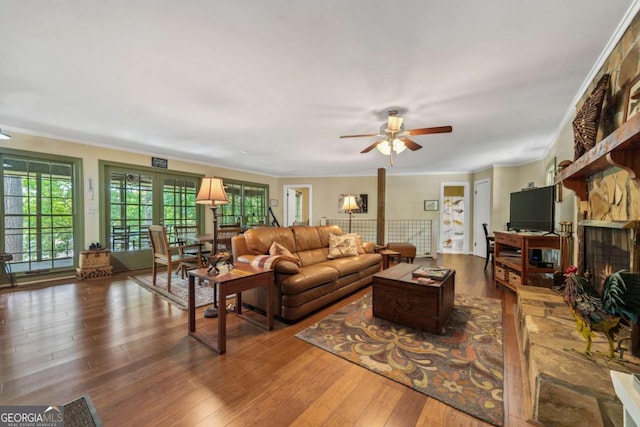 living area featuring hardwood / wood-style flooring, a ceiling fan, a stone fireplace, and ornamental molding