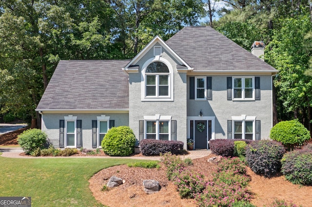 view of front facade featuring a chimney, stucco siding, a front yard, and a shingled roof